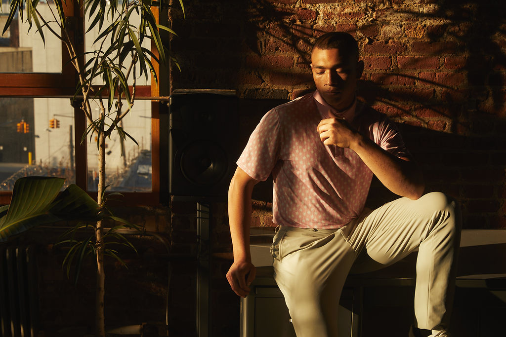 A man in a dimly lit room sitting on a table in front of a brick wall wearing a Sustainable Men's Pink Palm Polo Shirt Classic Fit and State of Matter khaki pants.