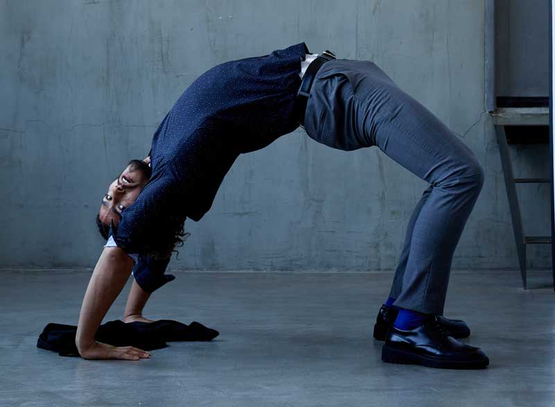 Man performing a backbend pose against a concrete backdrop, wearing State of matter's navy blue button-down T-shirt, gray chino pants, and  COOLMAX performance suit.