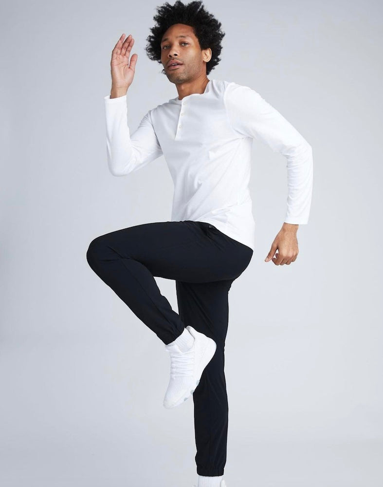 A man jumping on one leg wearing a State of Matter white Henley shirt and sustainable black joggers. 