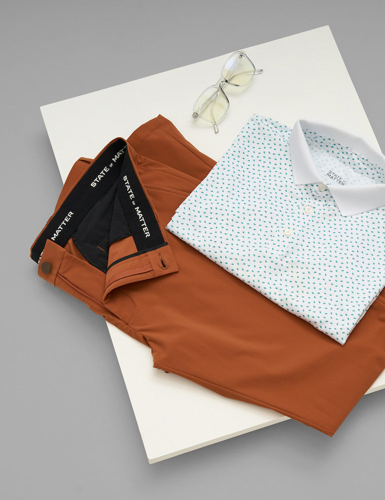 A table showcasing state of matter's mens sustainable orange chino pants and white polo shirt with green leaf print