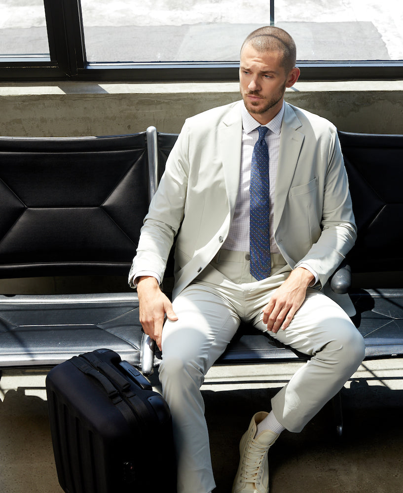 A man sitting in what appears to be an airport with a black suitcase wearing sustainable light beige business casual pants and a State of Matter light grey suit jacket with a light grey long sleeve button up and navy tie.   