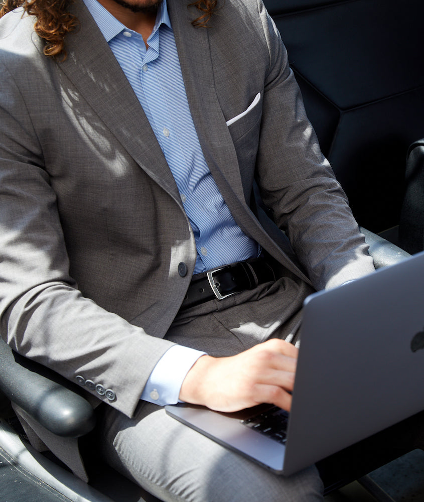 Man with curly hair sitting in a car, typing on a laptop, dressed in State of matter's gray COOLMAX performance suit paired with a light blue shirt and black belt.