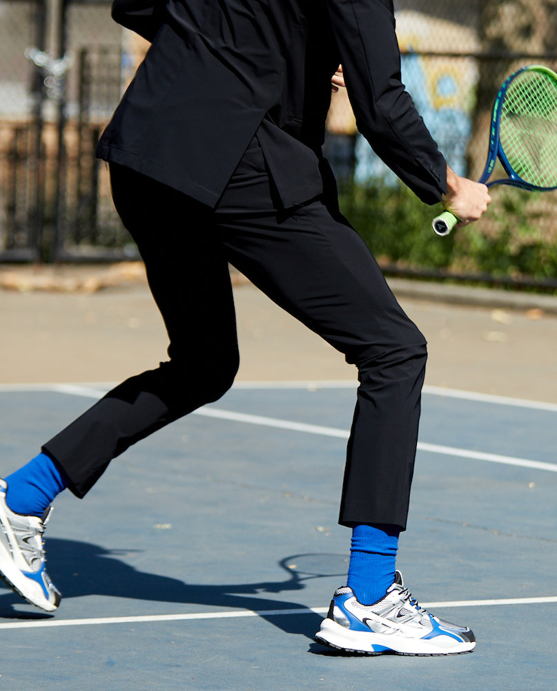 Image of the bottom half of a man playing tennis wearing State of Matter black chino pants and sustainable black men's suit jacket. 