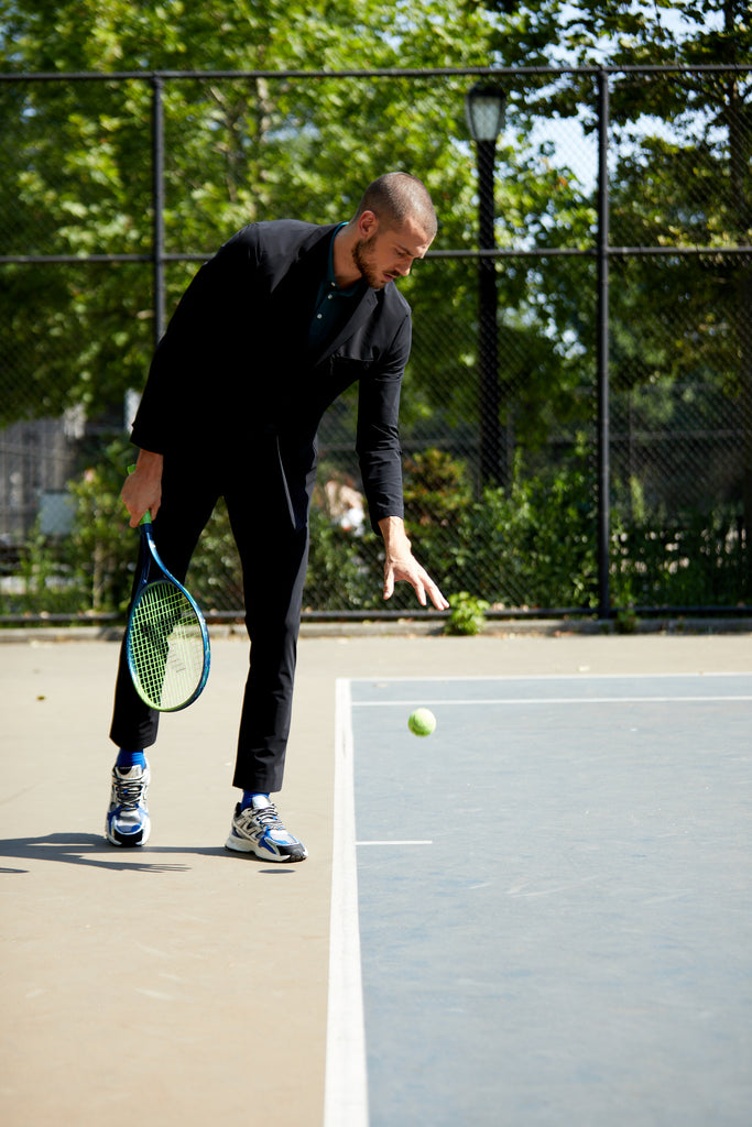 A man playing tennis on a tennis court with a racket in one hand and bouncing a tennis ball with the other. He is wearing a men's black suit jacket and State of Matter black chino pants with a sustainable black Henley shirt