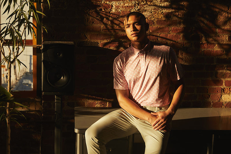 A man in a dimly lit room sitting on a table in a State of Matter Men's Pink Polo and sustainable chino khaki pants 