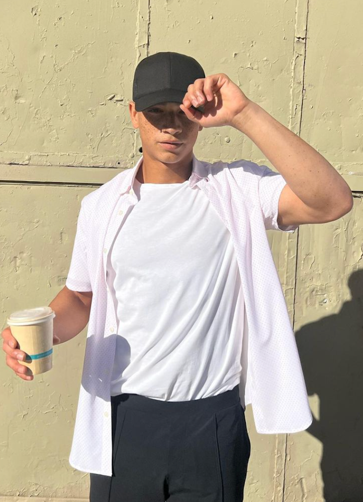 A man holding a cup of coffee in one hand and holding the rim of his black baseball cap. Wearing an open sustainable short sleeve button down 