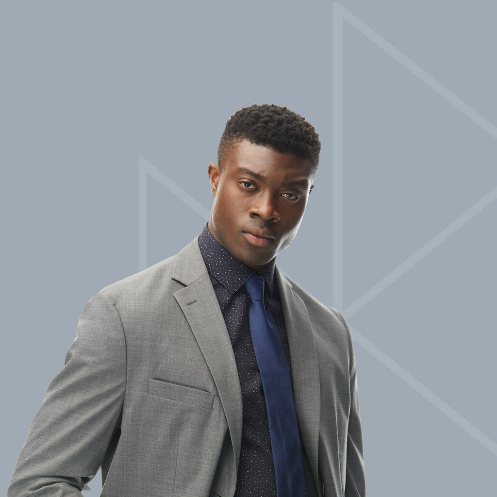 A man standing against the State of Matter logo wearing a Grey Cooling Performance Suit Jacket by State of Matter and Sustainable Men's Star Gaze Long Sleeve Dress Shirt with a navy tie