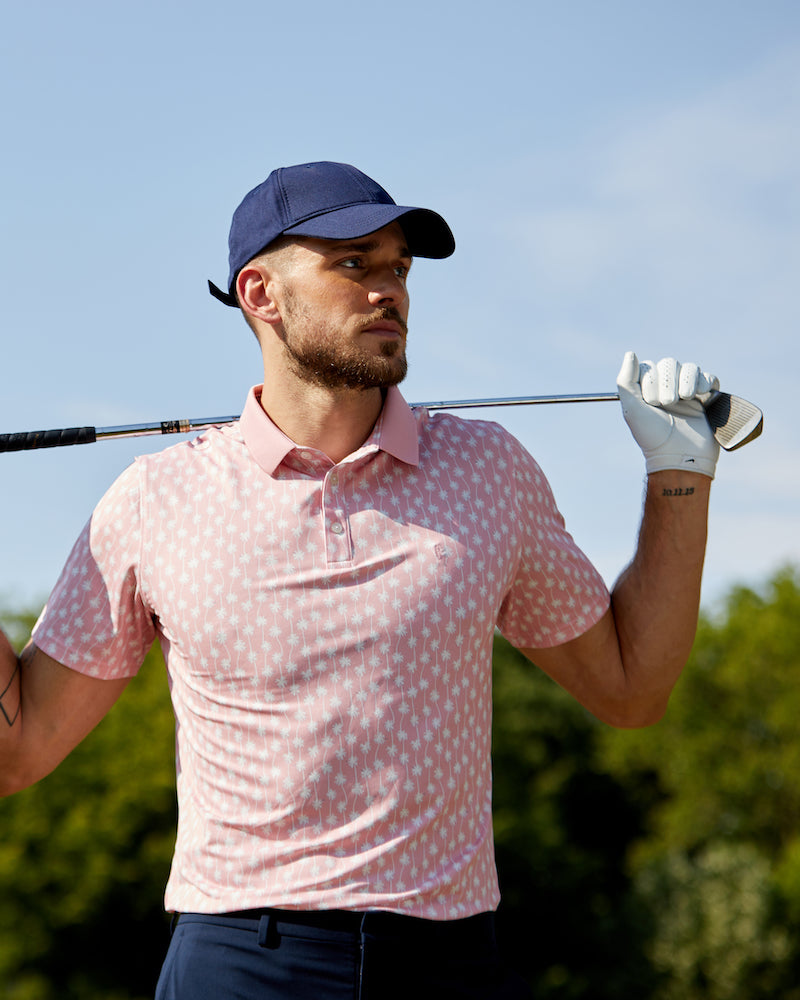 A man holding a golf club across his shoulders and wearing a State of Matter Sustainable Men's Pink Palm Polo Shirt Classic Fit.