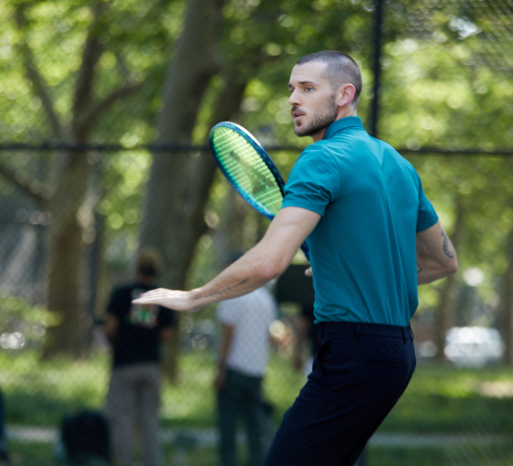 Man playing tennis while wearing state of matter sustainable men's turquoise polo and black chino pants