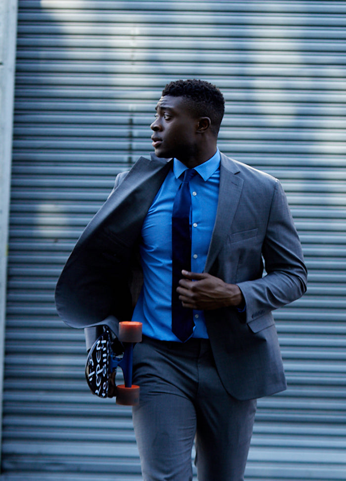A professional Black man in a tailored State of matter gray COOLMAX® suit and blue shirt carries a skateboard. He's glancing to the side, possibly signaling a moment of contemplation or observation. The corrugated metal background suggests an urban setting. This image could be used to illustrate the blend of business attire with an active sustainable lifestyle.